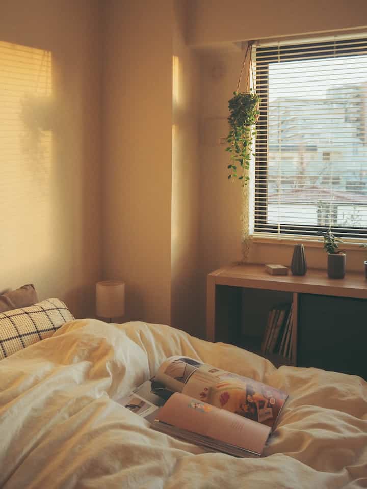 暖かな黄金色の光が差し込む、白とナチュラルカラーの寝室。ベッド、木製の棚、植物が配置された雰囲気のあるインテリア