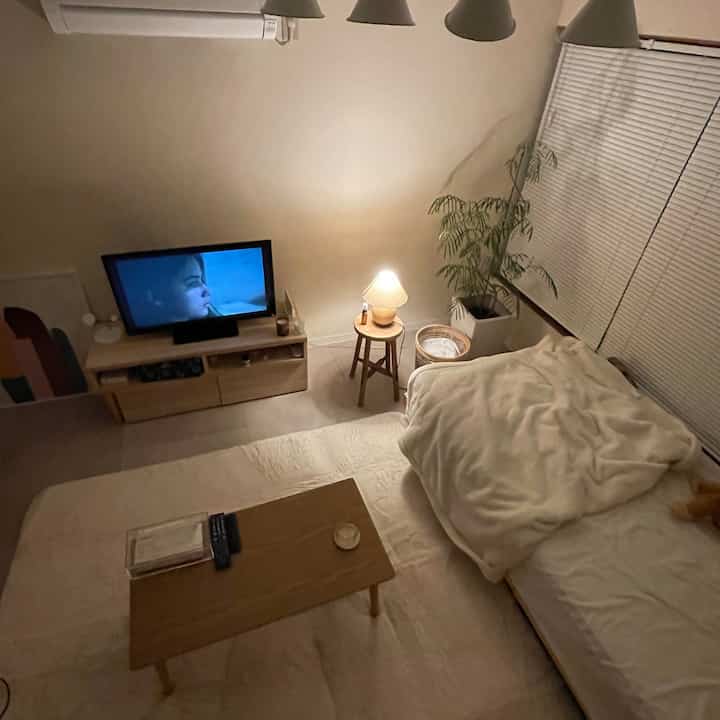 Soft white and wood-toned single household bedroom-living room featuring nightstand and table lamp with cozy atmosphere