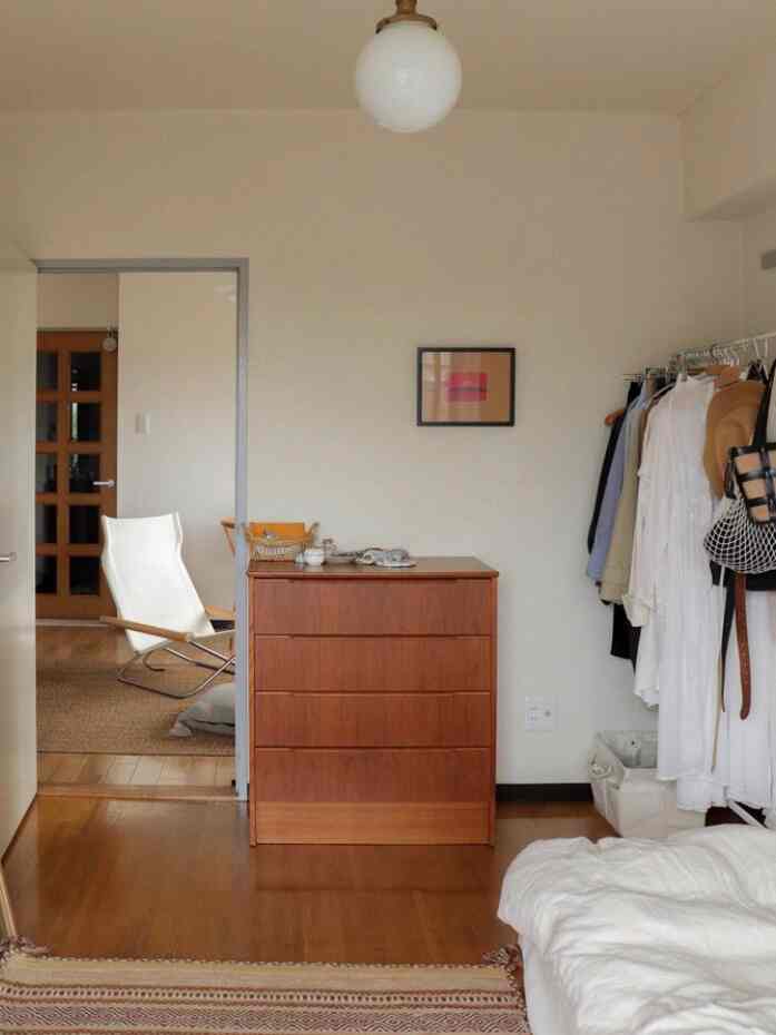 Natural tone bedroom featuring a wooden dresser, white bed, and clothing rack with a tidy and cozy atmosphere