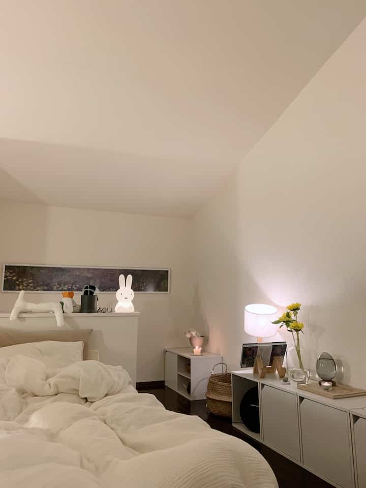 A white and beige toned bedroom featuring a cozy bed and storage units, creating a minimal and warm atmosphere