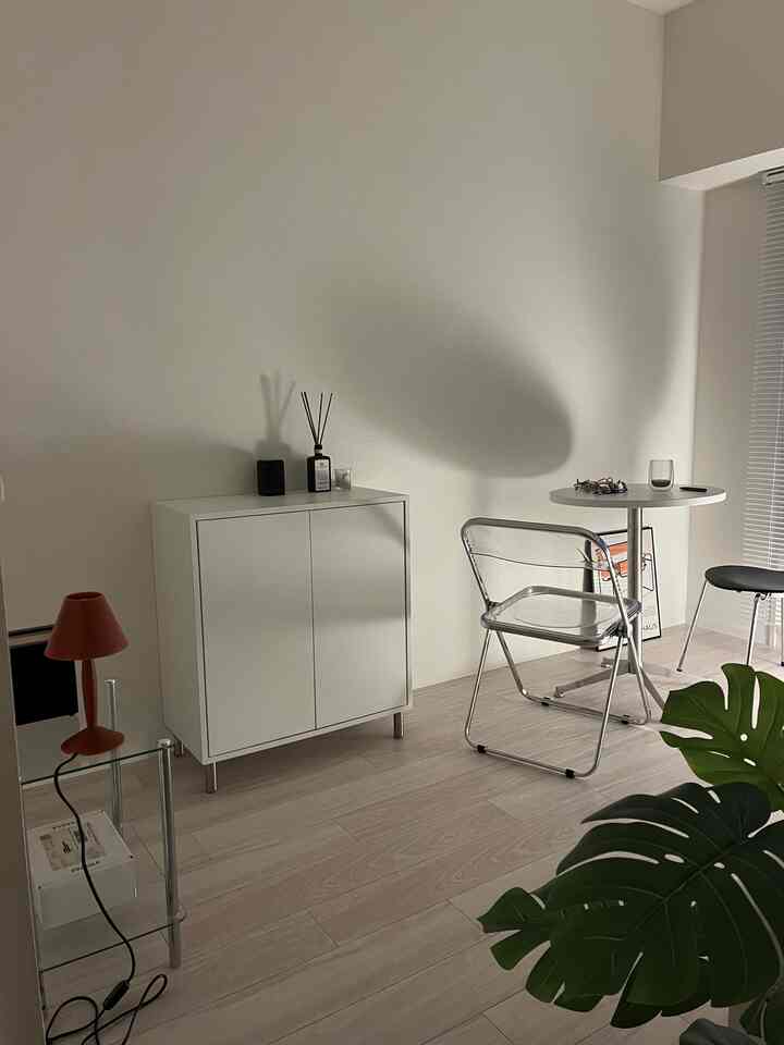 白を基調としたナチュラルトーンの8畳L字型の部屋、丸テーブルと透明チェア、赤いテーブルランプが配置されたシンプルな一人暮らし空間