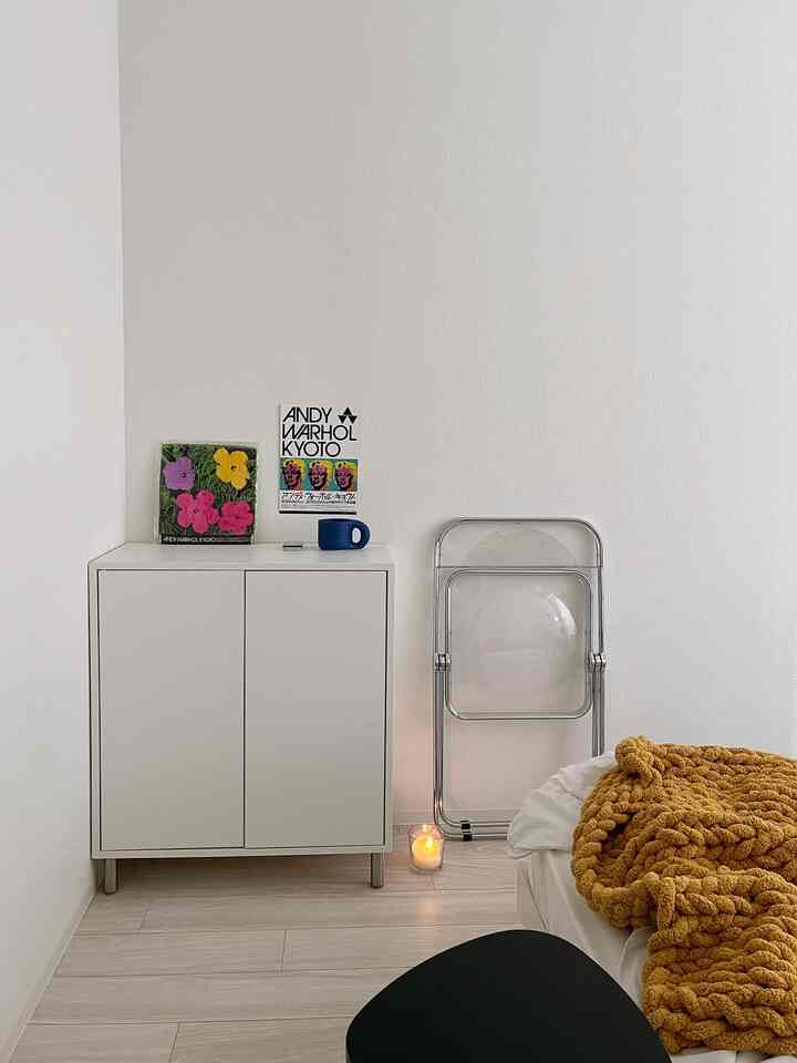 白い壁と床、シンプルな収納キャビネットが配置されたミニマルな寝室、マスタード色のブランケットがアクセント