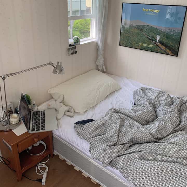 白とナチュラルトーンの寝室、チェック柄の掛け布団カバーとサイドテーブルに置かれたノートパソコンが配置された落ち着いた空間