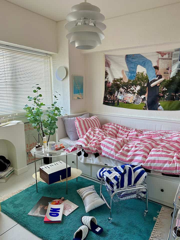 白とピンクのストライプのベッドカバーがある寝室、青いラグと植物が配置された落ち着いたナチュラルモダンな空間