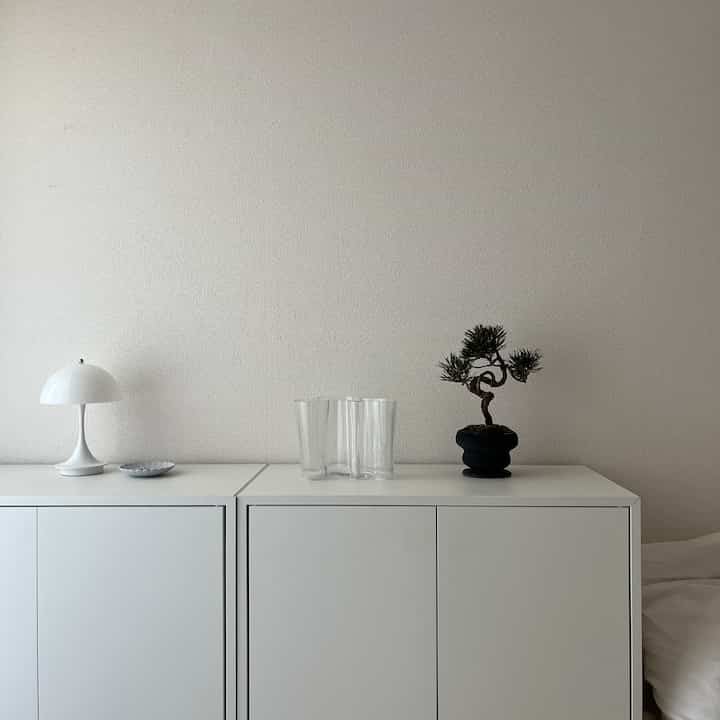 Bright white-toned studio apartment space featuring white shelves with a table lamp, transparent vase, and small bonsai plant, simple and natural interior style