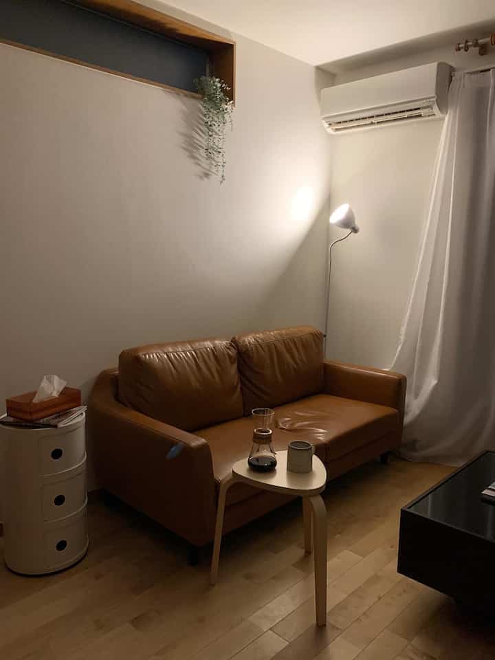 A white and brown toned studio apartment living room featuring a brown leather sofa, wooden stool, and floor lamp creating a cozy atmosphere
