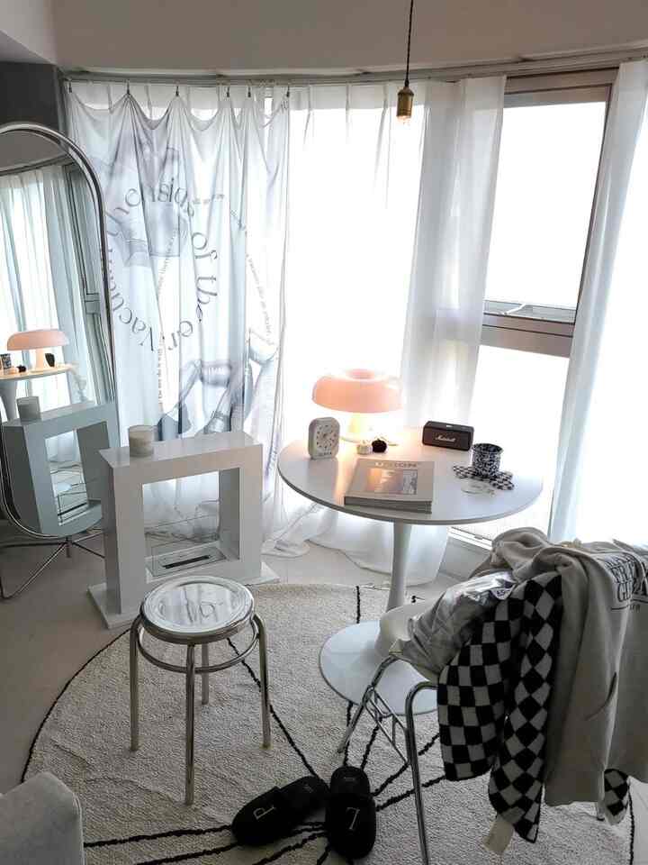 Bright white-toned living room space featuring a round table, silver stool, and large silver studio mirror with a minimal aesthetic