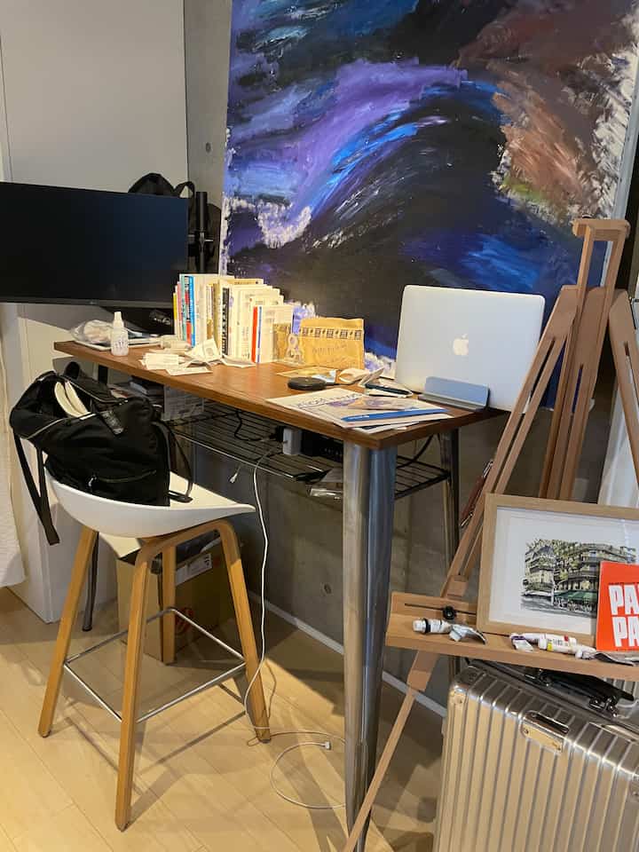 木目調とグレーの壁が調和するホームオフィス空間、高いテーブルとバースツール、本とノートパソコンが置かれたアートスタジオ風の作業スペース