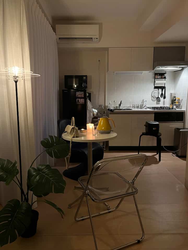 Cozy kitchen space with white curtains, natural toned cabinets, transparent stool, and plants under soft lighting