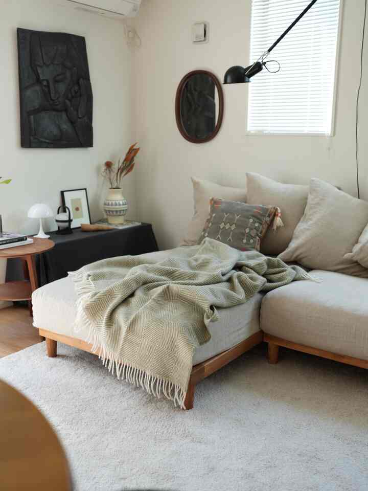 A soft beige and white toned living room featuring beige sofa with olive blanket, artwork on wall, and a black wall sconce.