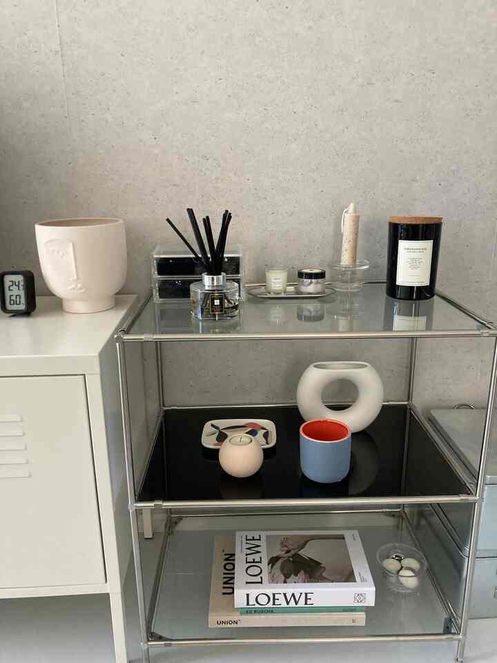 Modern corner storage rack against gray wall and white cabinet backdrop, featuring diffuser, candles, and decorative items with a clean atmosphere