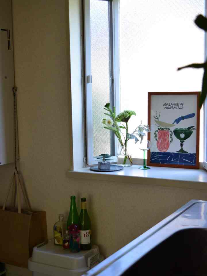 ベージュの壁と窓、キッチンカウンターのそばにポスターと植物が配置された温かみのあるキッチンスペース