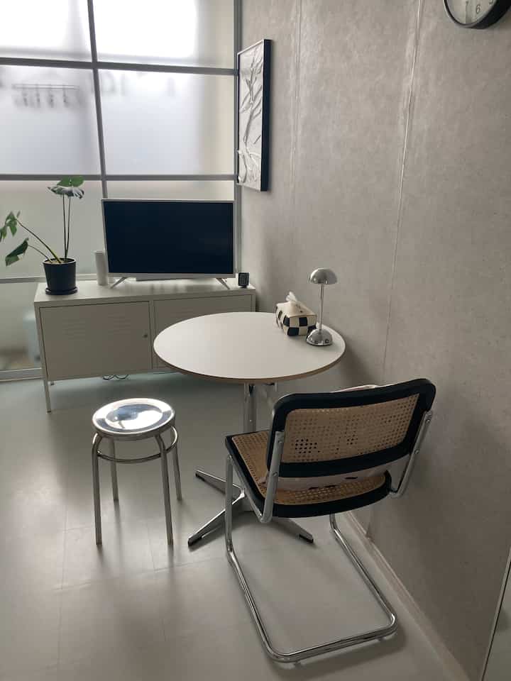 ライトグレーと白を基調とした狭い部屋に丸テーブルとラタンチェア、シルバースツール、テーブルランプが配置されたシンプルな空間