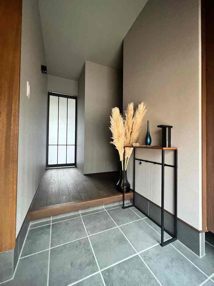 グレーのタイル床と木目調のフローリングが調和するナチュラルスタイルの玄関、コンソールテーブルとパンパスグラスの花瓶が配置された空間