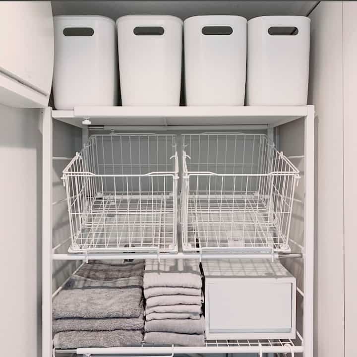 白を基調としたコンパクトなバスルーム、白い収納ラックとバスケットが配置され、整頓されたタオルがある実用的な空間