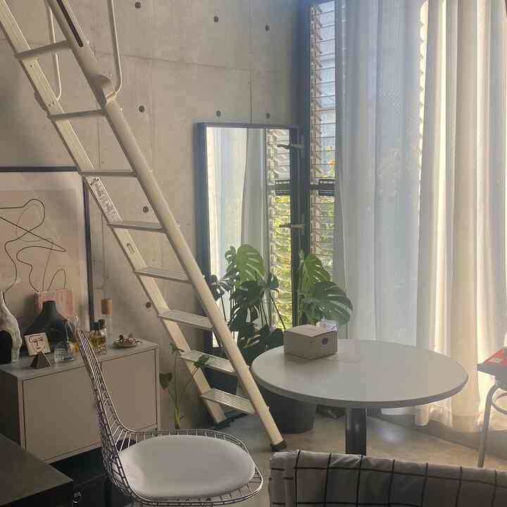 グレーのコンクリート壁と白いダイニングテーブルがあるナチュラルモダンのリビング、窓際に大きな鏡と植物が配置された印象的な空間