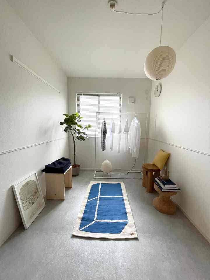 ナチュラルな色調の縦長で小さな部屋、中央に青と白のパターンラグが置かれ、ハンガーラックと植物が調和したシンプルな空間