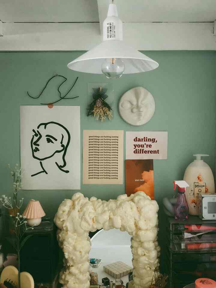自然なライトグリーンの壁と白いペンダントライト、ユニークな泡のフレームミラー、多様なウォールアートが配置されたミニマルなホームオフィス空間