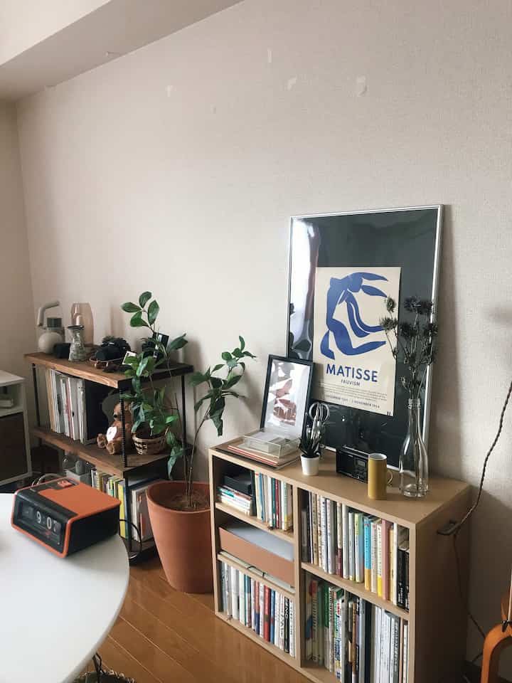 Natural toned small living room featuring wooden shelves and bookshelf with a large Matisse poster leaning against the wall, cozy atmosphere