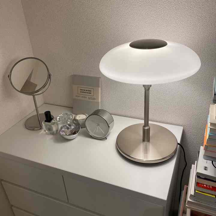 A clean space featuring a white chest of drawers with a silver-based table lamp, mirror, alarm clock, and candles arranged on top