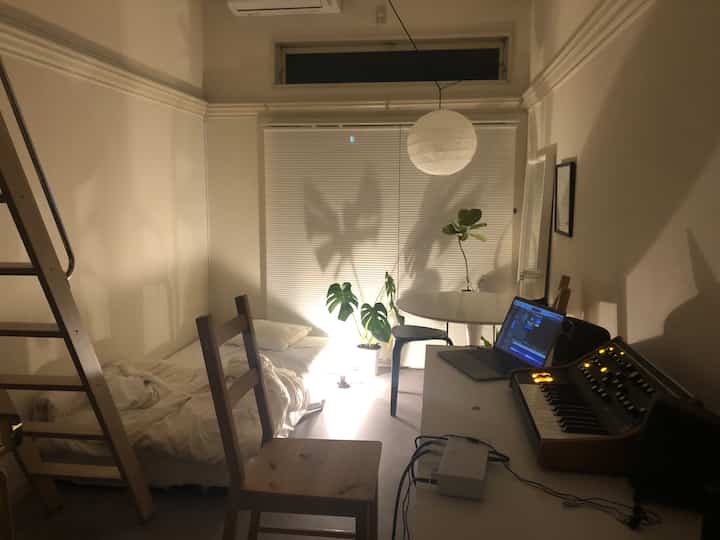 温かみのある木目調と自然光が調和した寝室、音楽作業用デスクとシンプルな家具が配置された雰囲気のある空間