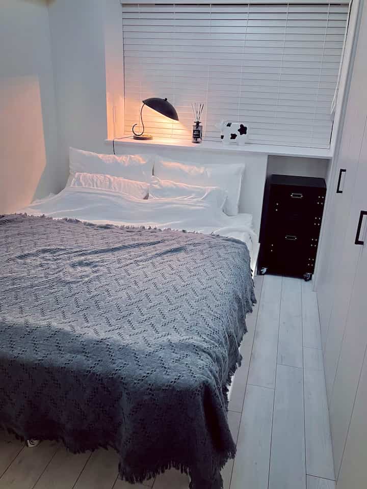 モノトーンの寝室、白いベッドとグレーのブランケット、黒い金属製ランプとチェストが配置されたシンプルで落ち着いたインテリア