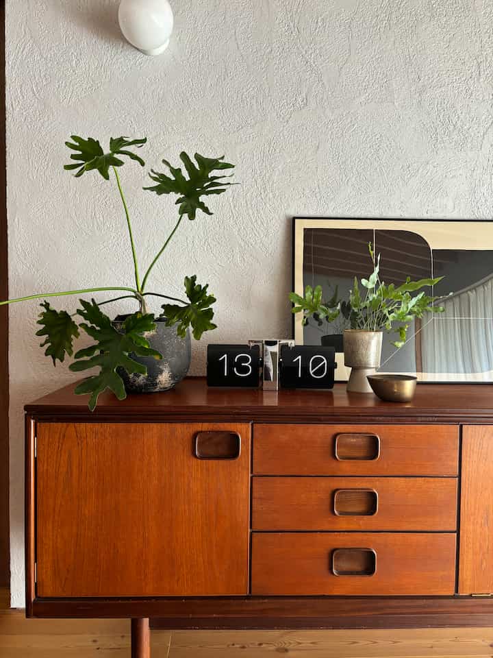 Natural style interior with a brown wood tone sideboard topped with two plants and a black flip clock against a textured white wall