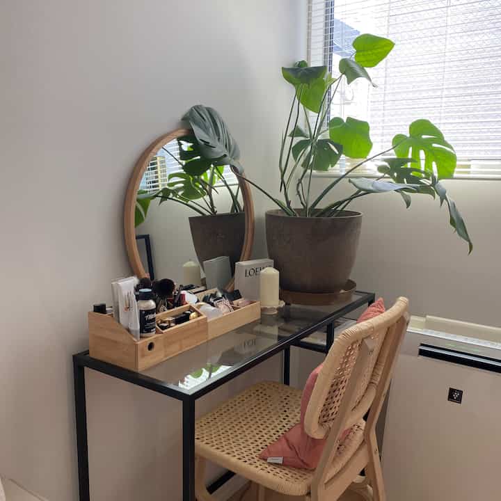 Bright white-walled small room featuring a wood-toned vanity with plant and rattan chair, presenting a minimal aesthetic
