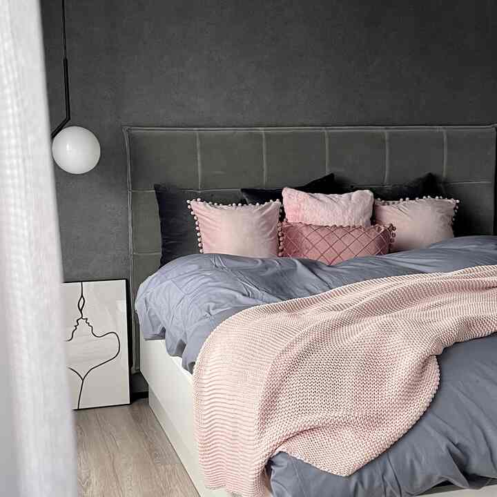 グレーの壁にピンクのクッションが配された寝室。白いベッドフレームとブランケットがあり、シンプルでモダンな雰囲気のインテリア