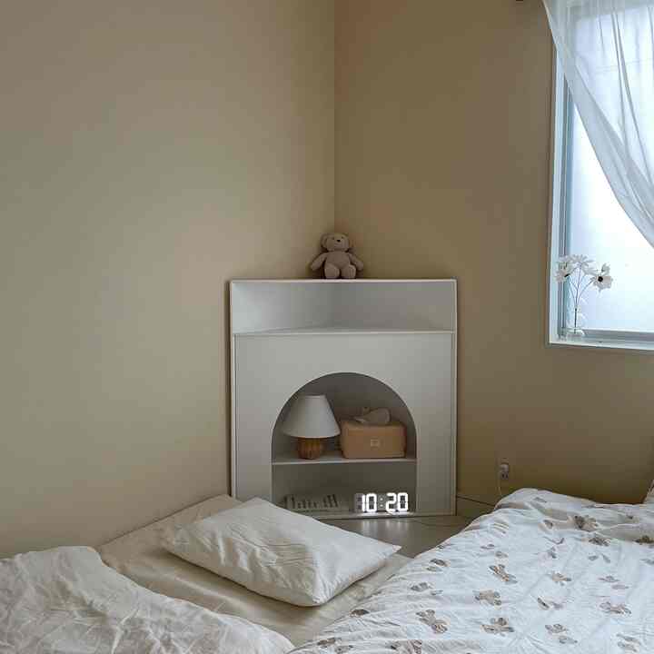 ホワイトとベージュの色調の寝室、コーナーに配置されたホワイトのコーナー収納とベッドがある落ち着いた空間