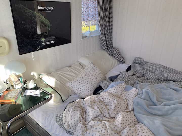 白と青のトーンの寝室、中央右にベッドと寝具、左にナイトテーブルとランプが配置された落ち着いた空間