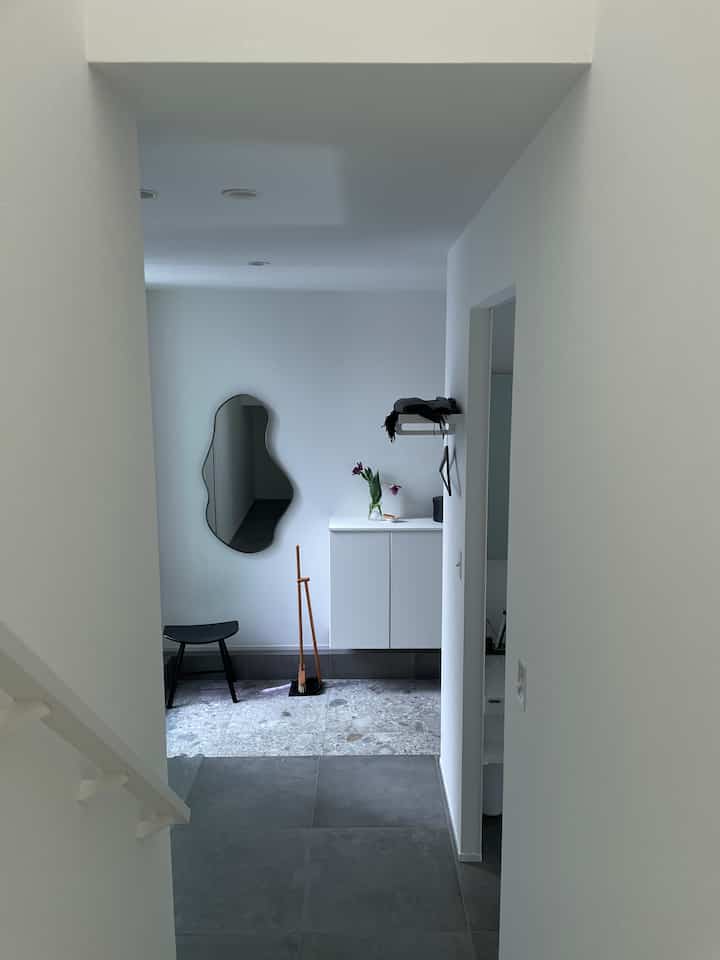 ホワイトとグレーのトーンが調和した玄関、独特な形の鏡とスツール、花瓶が配置された清潔感のあるモダンな空間