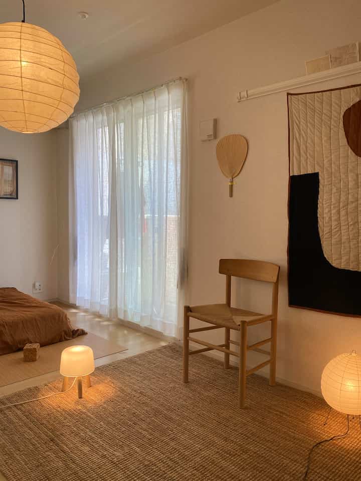 暖かいベージュとナチュラルトーンのリビング、木製の椅子とラタンラグ、多彩な照明が配置された雰囲気のあるインテリア