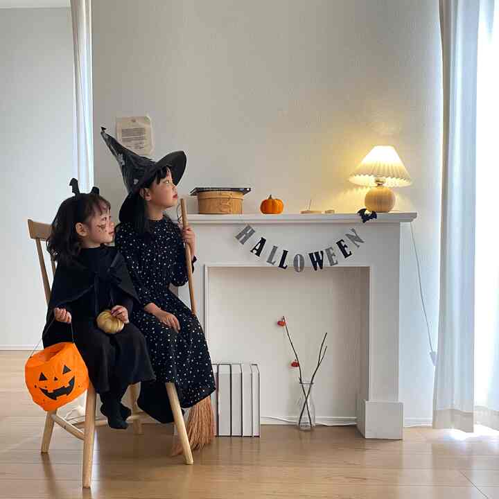 明るいホワイトの壁と木目調の床がある子供部屋で、ハロウィン装飾とテーブルランプが配置された雰囲気のあるインテリアに、2人の子供が椅子に座っている