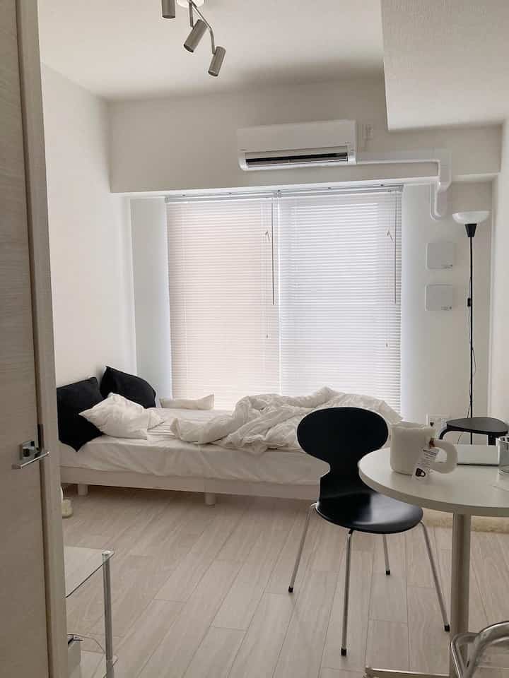 White and light brown toned compact studio bedroom featuring a bed, blinds, and a black armchair in a minimalist setting