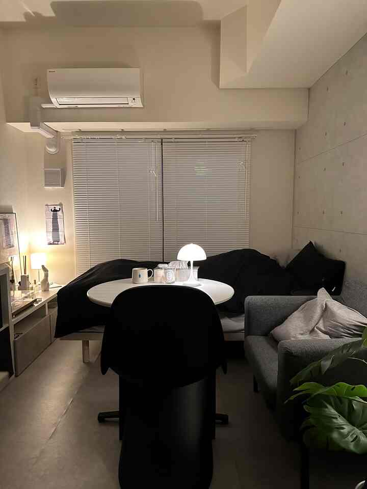 A cozy living room in gray and black tones featuring a window with blinds, a white round table under warm lighting, and a gray armchair