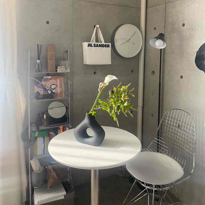 Light gray concrete wall and white round cafe table in a small room with metal wire chair and floral vase, natural modern interior