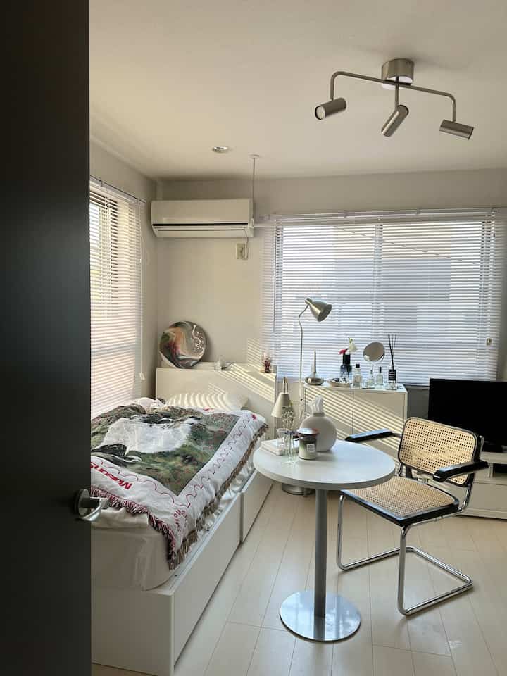 Small bedroom in white and light brown tones, featuring a modern bed, rattan chair, and round table with a calm atmosphere