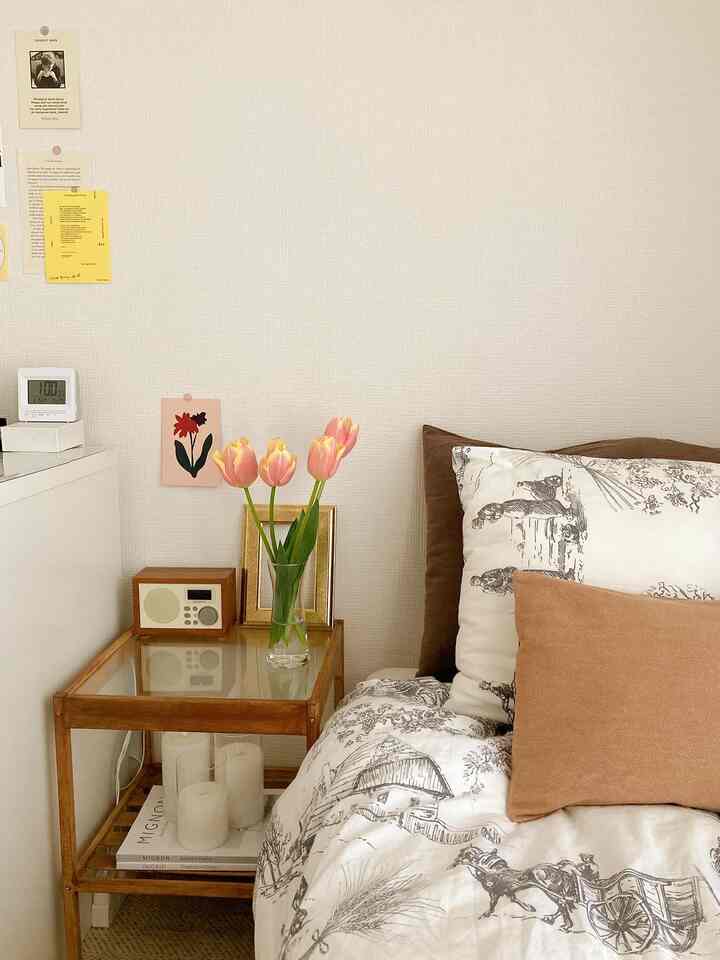 ナチュラルなトーンの寝室、ナイトテーブルの上にピンクのチューリップの花瓶とゴールドの額縁が置かれた雰囲気のあるインテリア