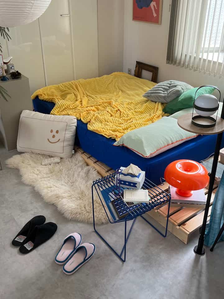 Natural-toned bedroom featuring bright yellow blanket and blue bedding, simple side table and soft rug arranged neatly