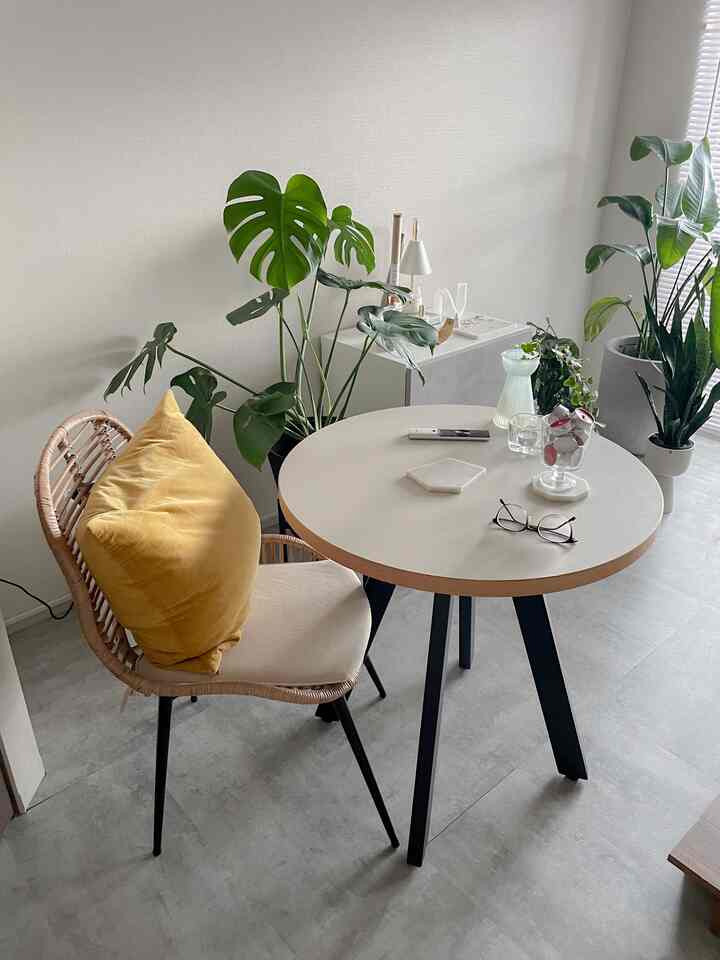 Natural-toned 6-tatami room featuring a round coffee table with black legs, rattan chair with yellow cushion, and various indoor plants in a cozy home cafe setup