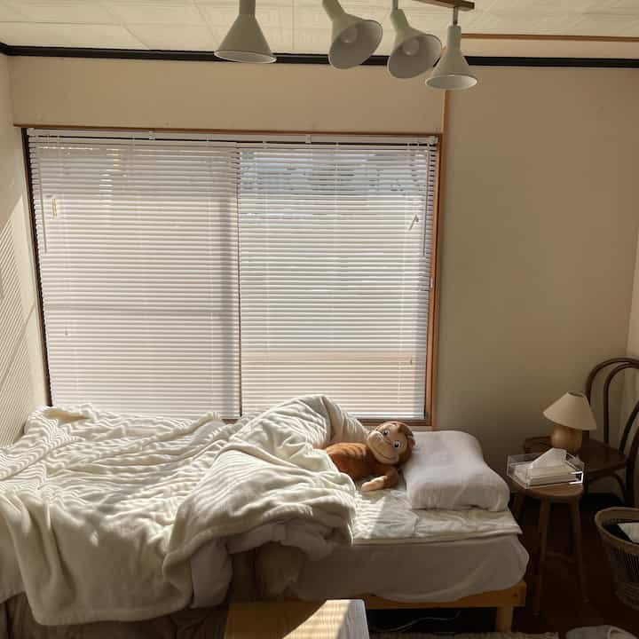 明るいベージュとナチュラルトーンの寝室、白いブラインドと毛布と枕が置かれたベッドがある雰囲気のある空間