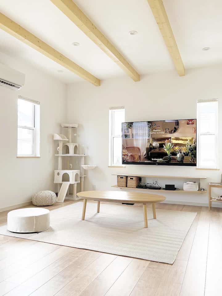 Bright white and natural wood tone living room featuring a central oval wooden table, large carpet, and a cat tower at the left corner in a simple cozy atmosphere