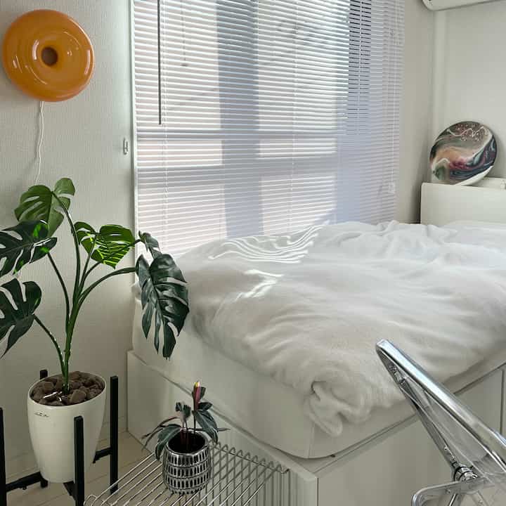 明るいホワイトトーンの6畳寝室、白いベッドとブラインド、左側に植物が配置されたシンプルな空間