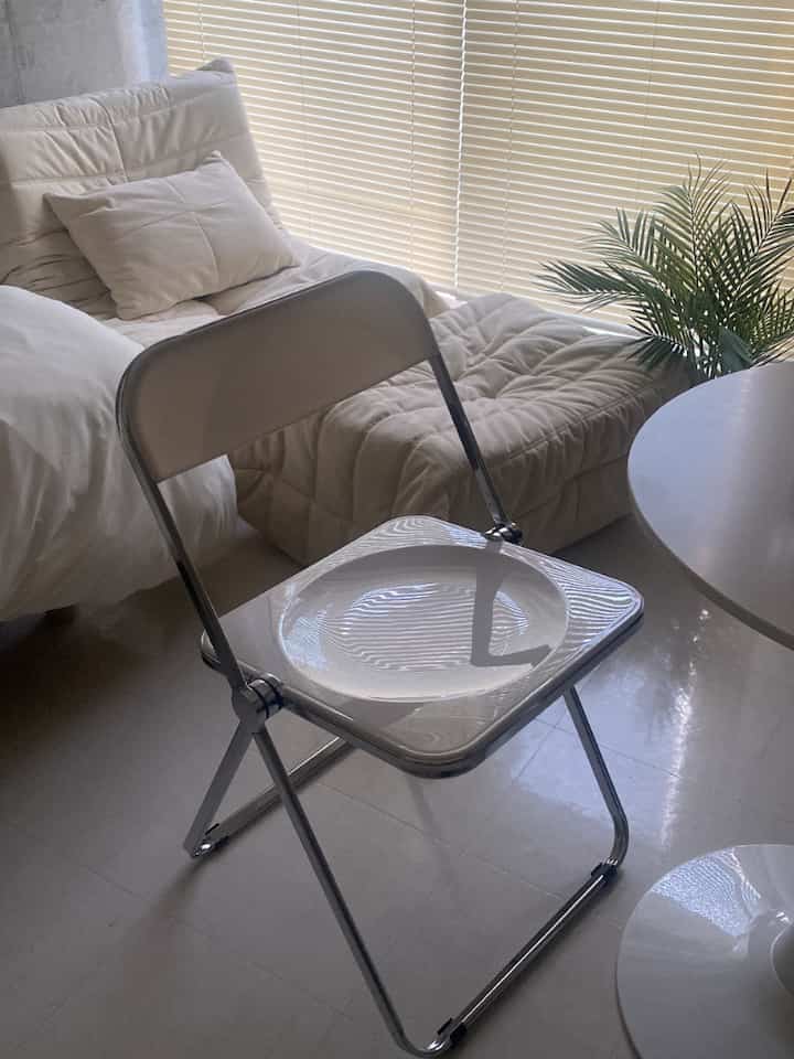 白とベージュのトーンのリビングルーム、折りたたみ椅子、クッション付きのソファ、丸テーブルと植物が配置された落ち着いた空間