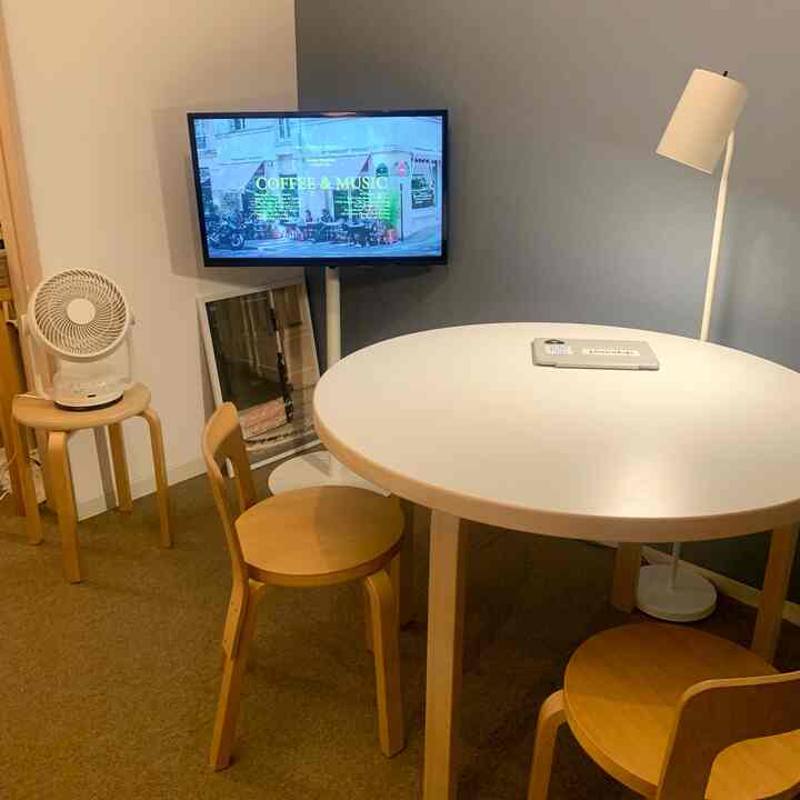 白色の丸テーブルとライトブラウンの椅子があるコンパクトなリビング、フロアランプとテレビ台が配置された雰囲気のあるインテリア