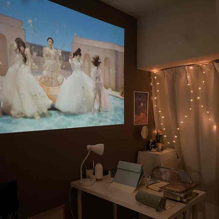Cozy small home office with brown wall and white curtains adorned with LED string lights, featuring a projector screen and a desk setup