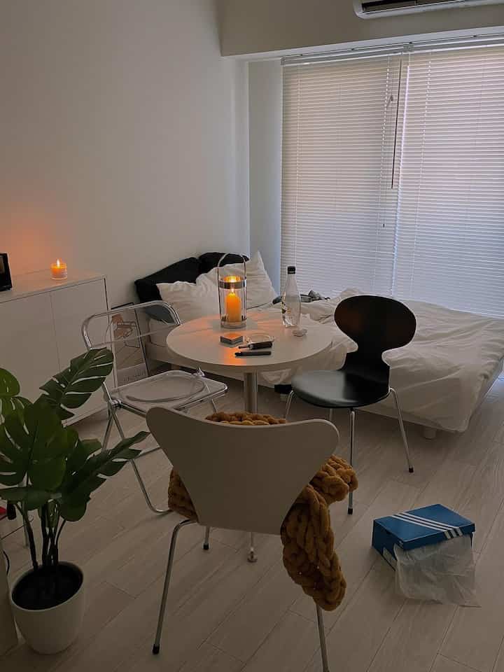 白い壁とブラインド、木目調の床が調和する一人暮らし用ワンルームの寝室兼ダイニング空間、中央のテーブル上のキャンドルが温かみのある雰囲気を演出