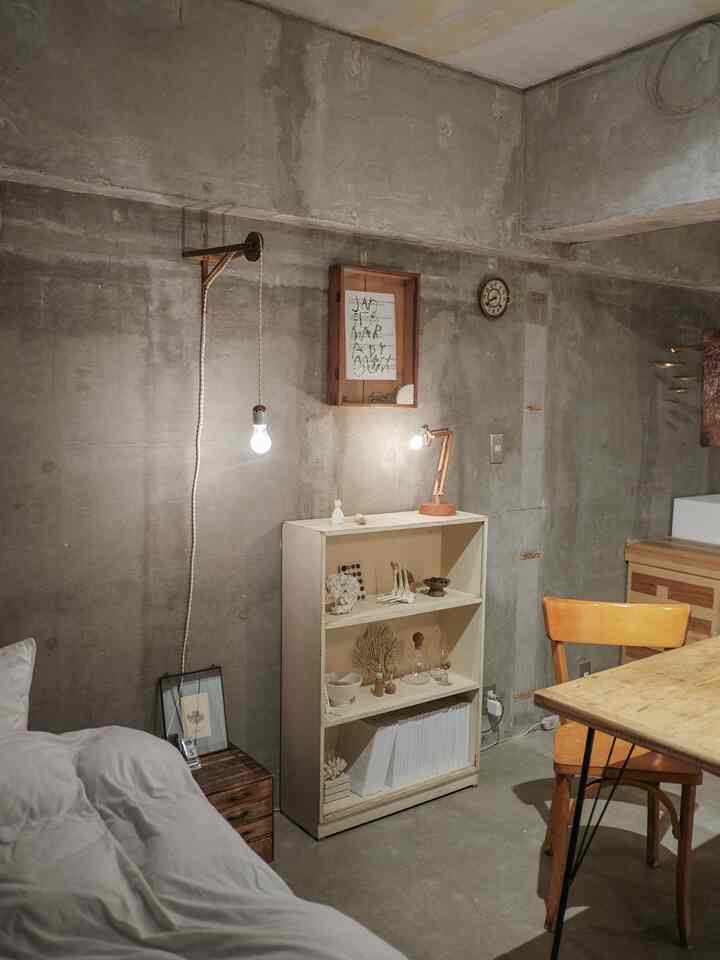 A vintage-style studio apartment bedroom featuring gray concrete walls, wooden furniture, a shelf, and ambient lighting