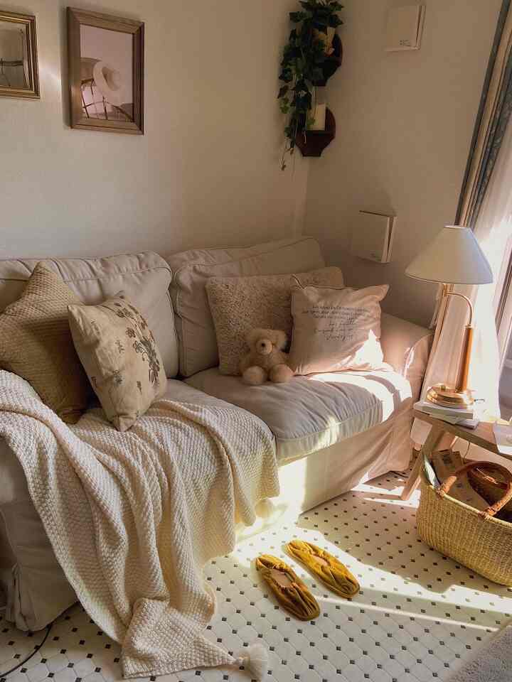 ベージュと白を基調としたリビング、ソファにブランケットとクッション、テディベア、テーブルランプが配置された温かみのある雰囲気のインテリア